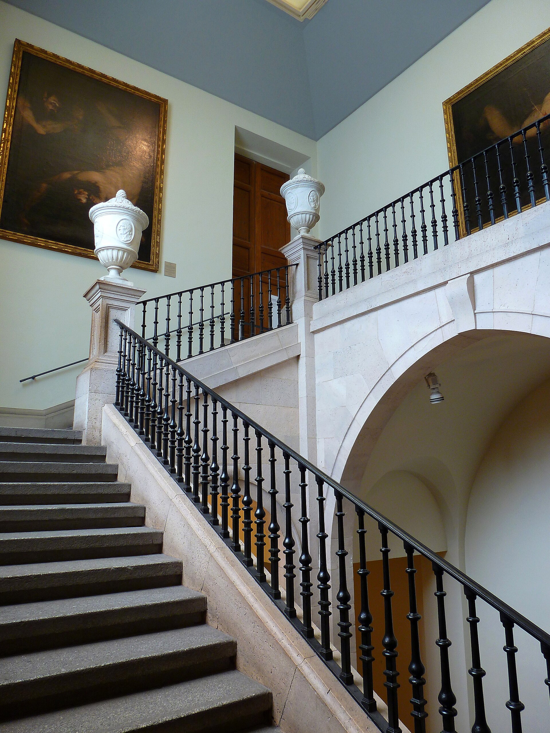 A stone staircase with a black iron balustrade, carved urns on pedestals and dark paintings on the landing walls