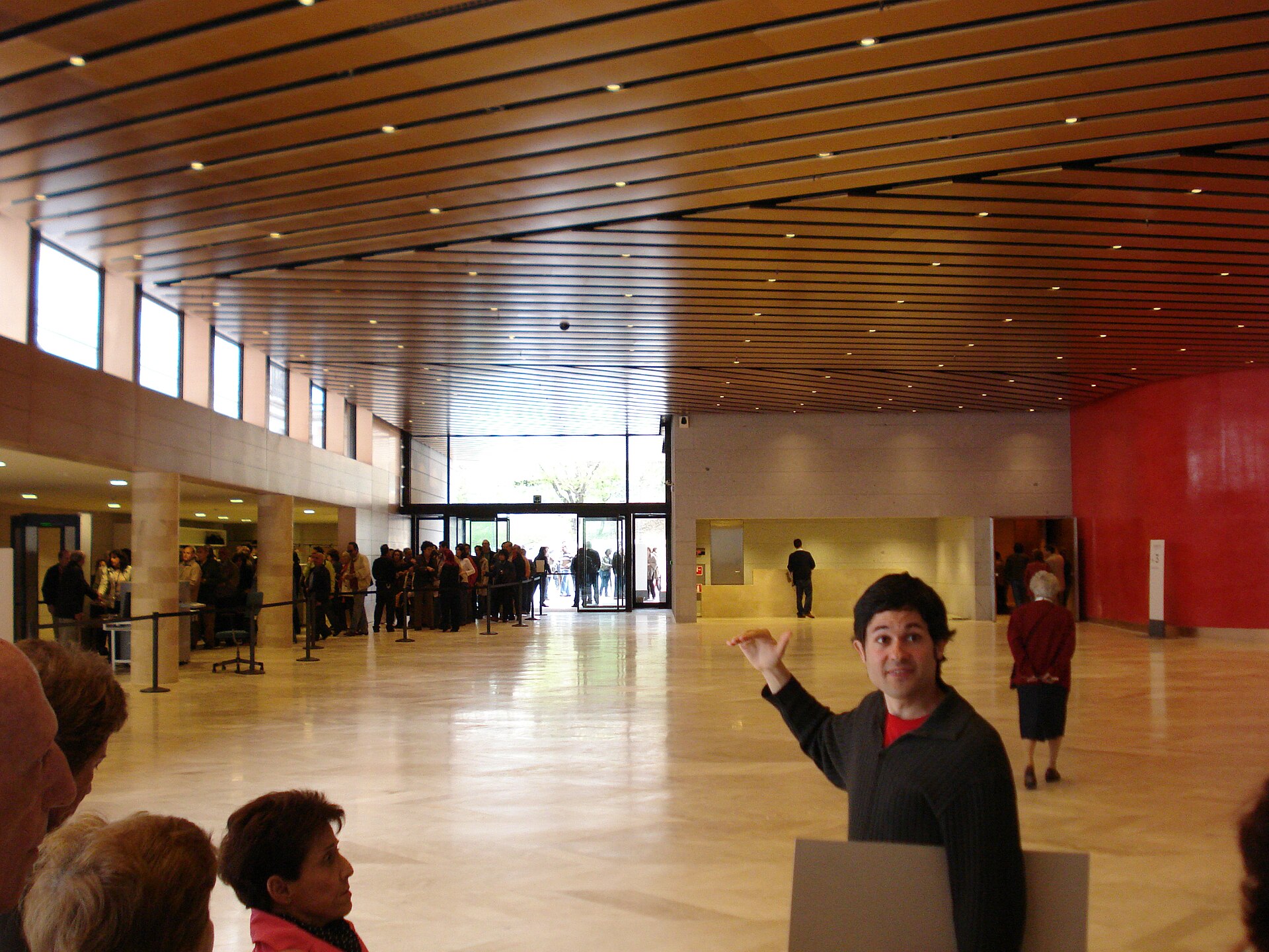 A wide entrance hall with a timber-slatted ceiling, pale stone floor, a red wall to one side and a ticket queue to the other
