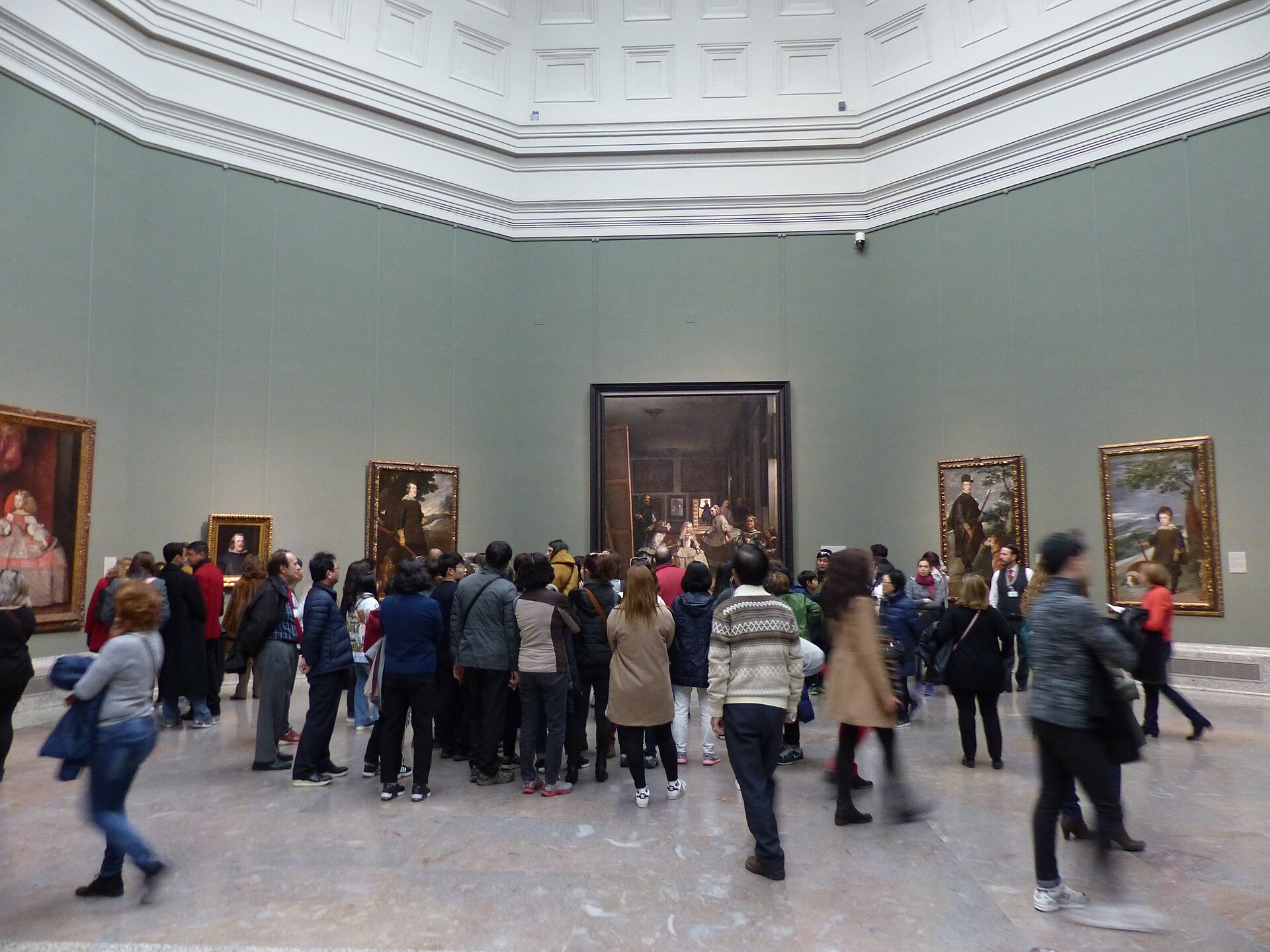 A grey-green room with Las Meninas on the end wall, ringed by Velazquez royal portraits, with a crowd in front of it