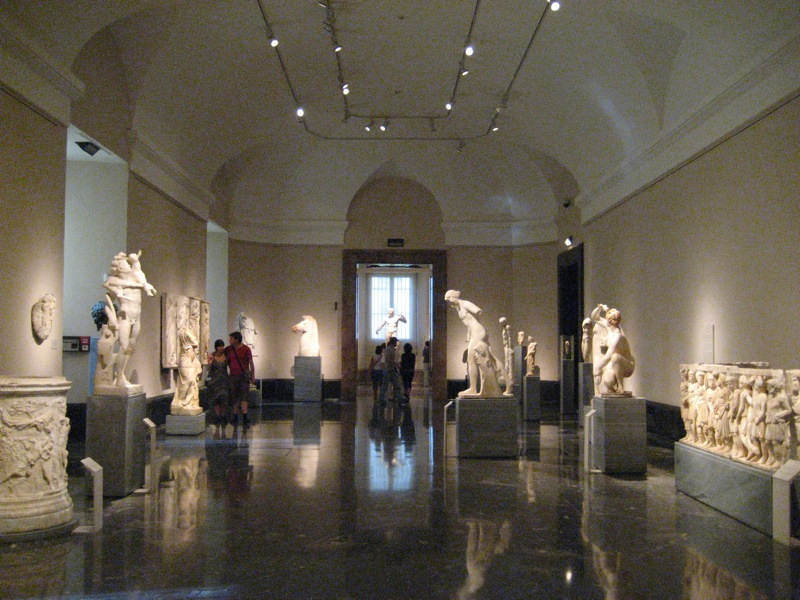 A ground-floor hall of marble statues on plinths with carved sarcophagus and puteal reliefs, polished dark floor