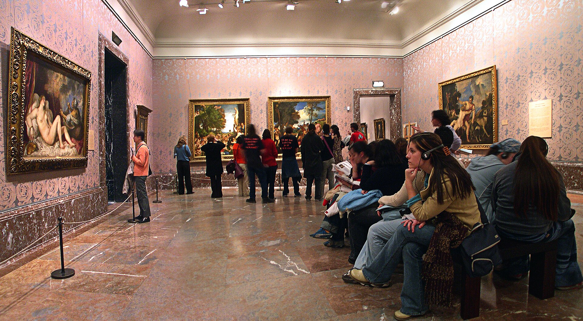 A room with patterned pink wall covering and marble dado hung with large Titian mythologies, visitors seated on a bench