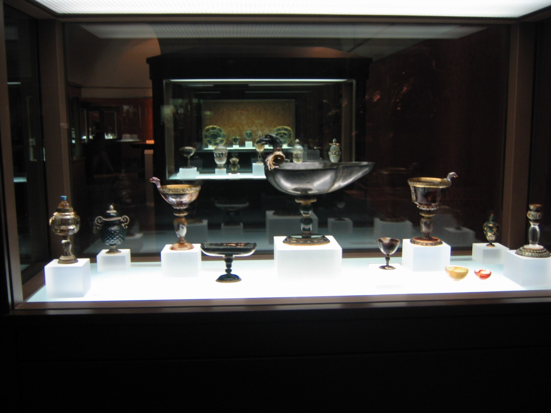 A lit vitrine of rock-crystal and hardstone vessels with gold mounts, a second case reflected behind