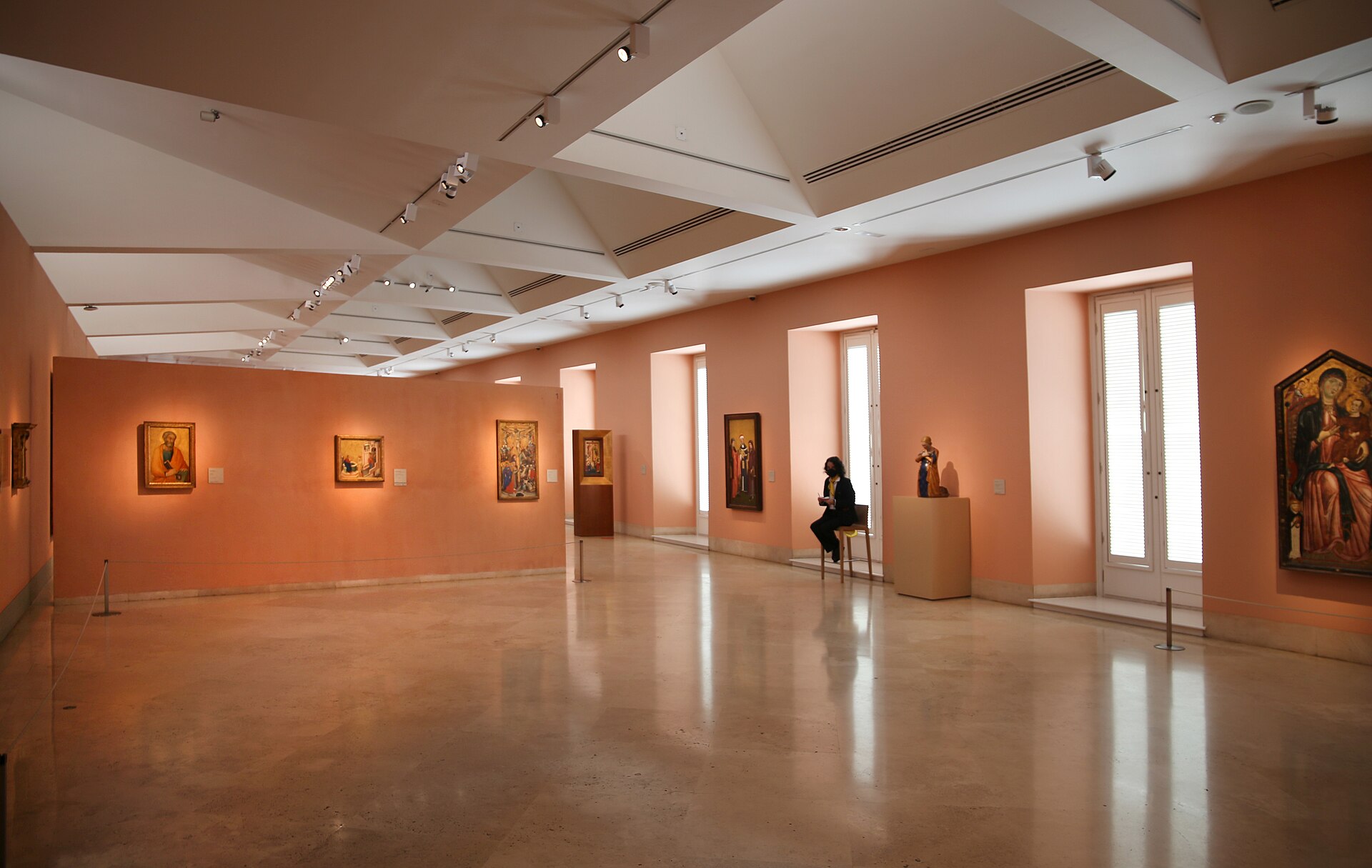 A top-lit gallery with terracotta walls hung with small gold-ground Italian panel paintings, a visitor seated on a central bench