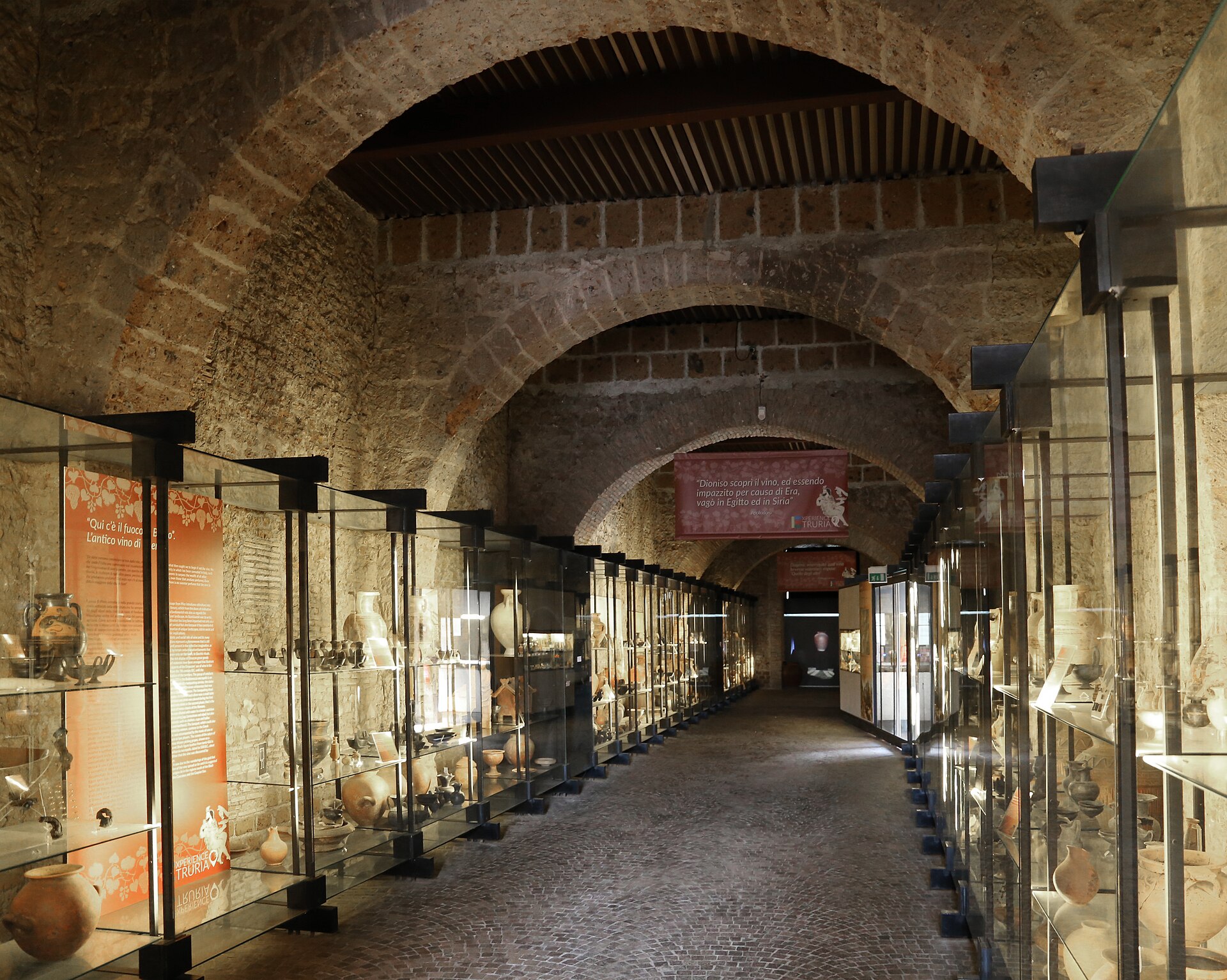 A vaulted stone and brick gallery inside the museum, lined with glass display cases of Etruscan pottery and grave goods