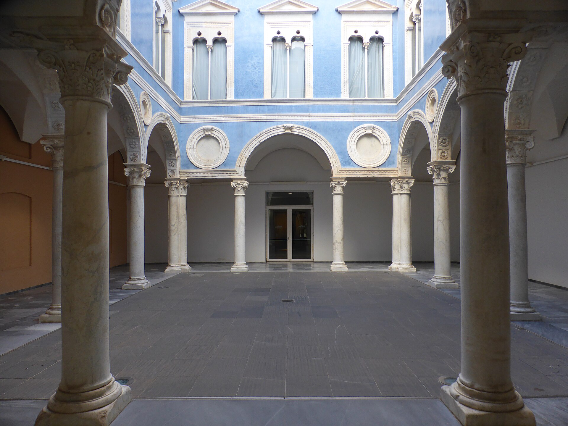 A two-storey Renaissance courtyard with paired marble columns, round arches and a blue upper facade with three arched windows, seen from inside the colonnade
