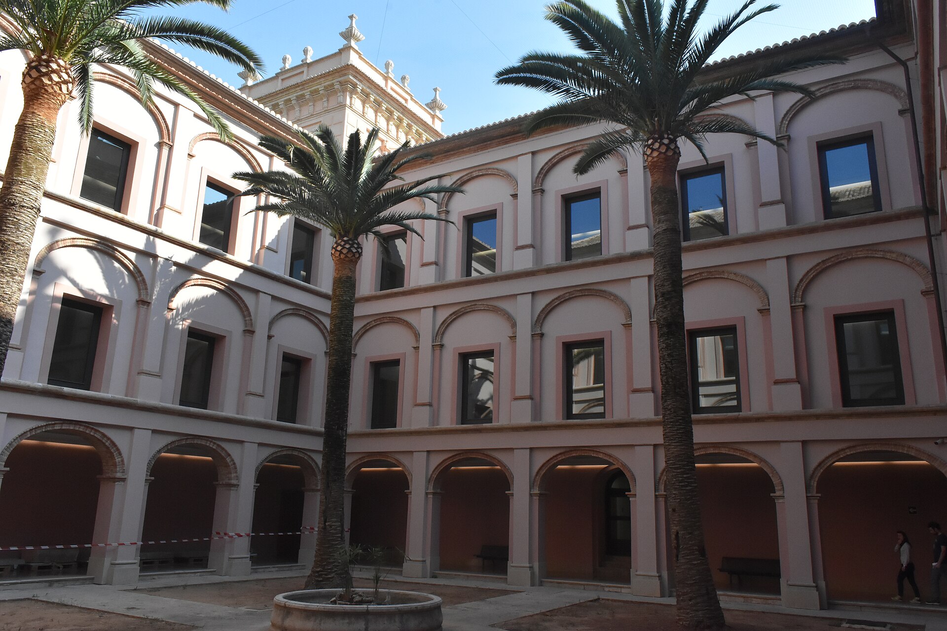 A renovated arcaded cloister courtyard with pale pink and cream walls, three storeys of arched windows, and tall palm trees planted at ground level