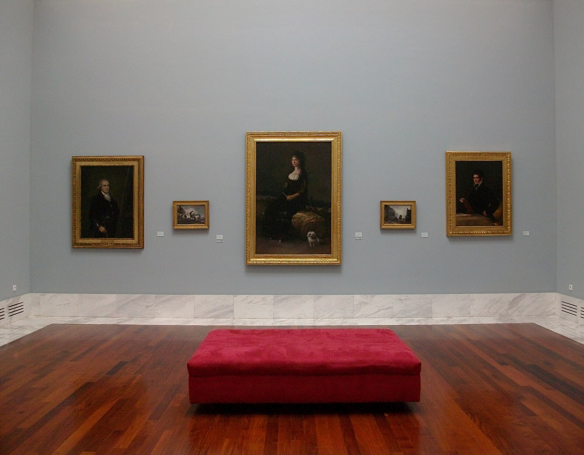 A museum gallery with pale blue-grey walls hung with gilt-framed portraits either side of a large full-length portrait, a red bench at the centre of the room