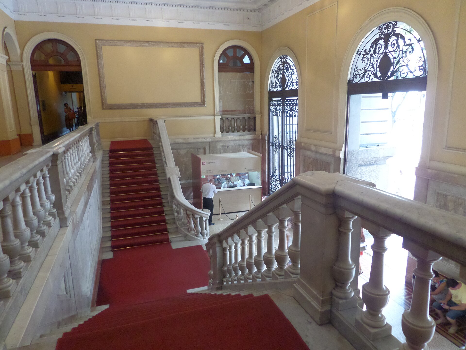 A double marble staircase with a red carpet runner and ornate stone and iron balustrades, viewed from an upper landing looking down toward arched glass doors