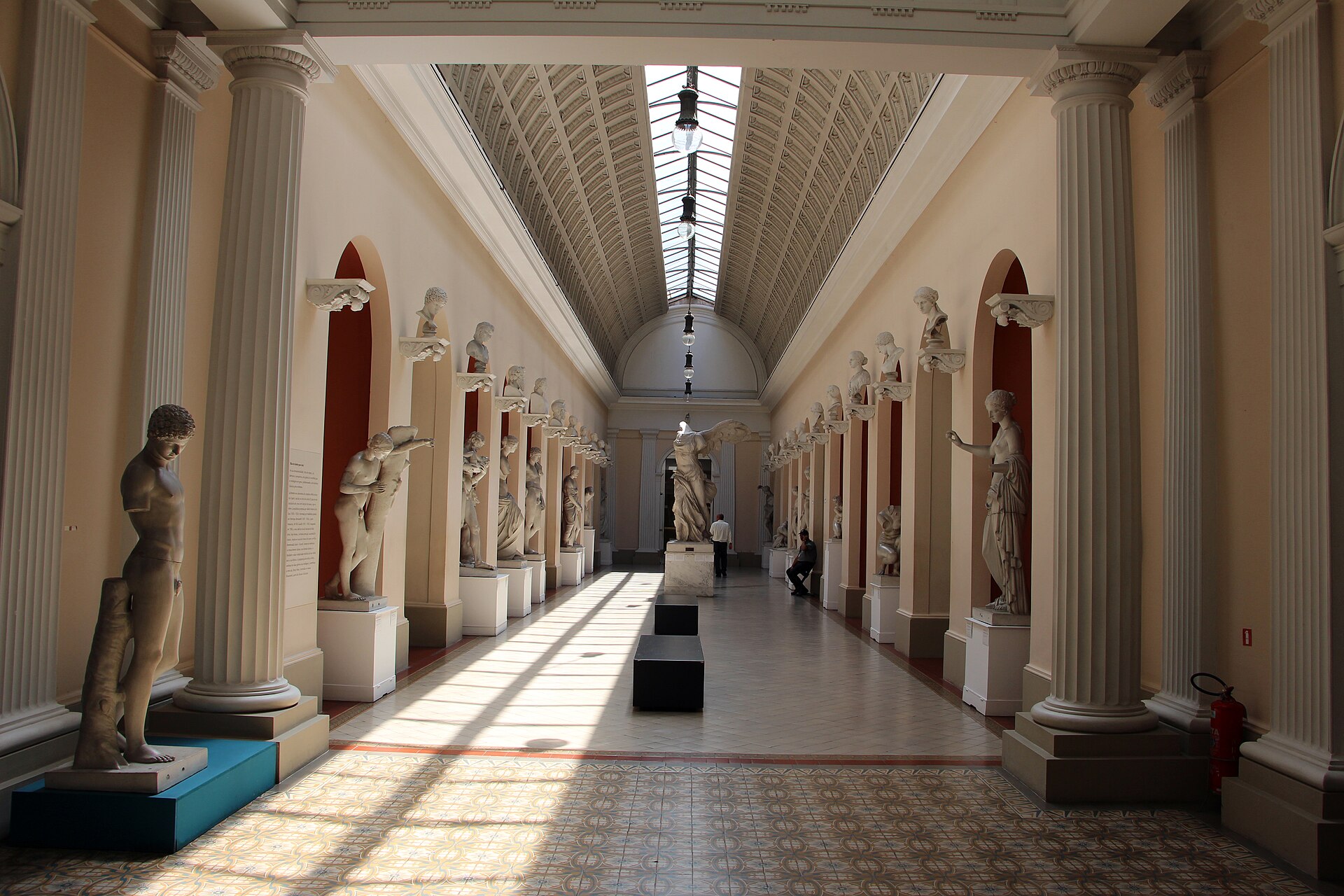 A long barrel-vaulted gallery lined with plaster cast sculptures of classical figures on pedestals under rows of skylights, with visitors walking between them