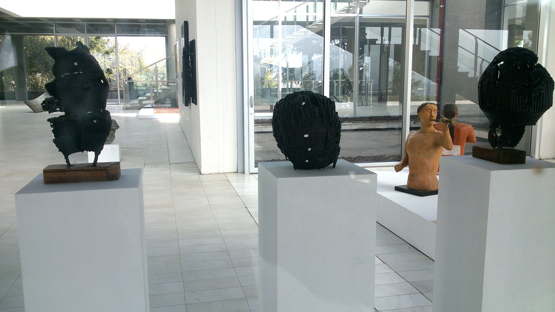 Terracotta figures and abstract stone sculptures displayed on plinths in the museum's glazed ground-floor foyer, with the outdoor sculpture court visible through the glass wall behind them
