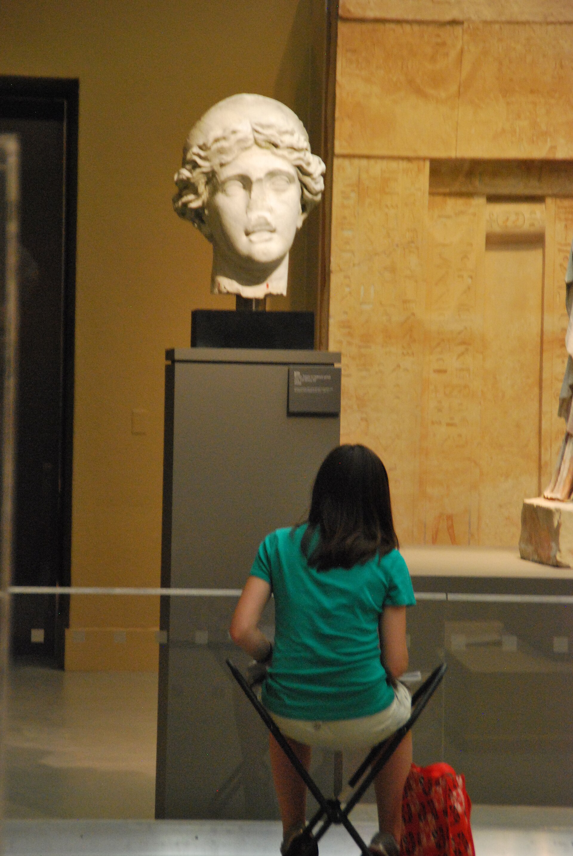 A visitor seated before a Greco-Roman marble head, with a hieroglyph-carved panel visible in the Egyptian galleries beyond