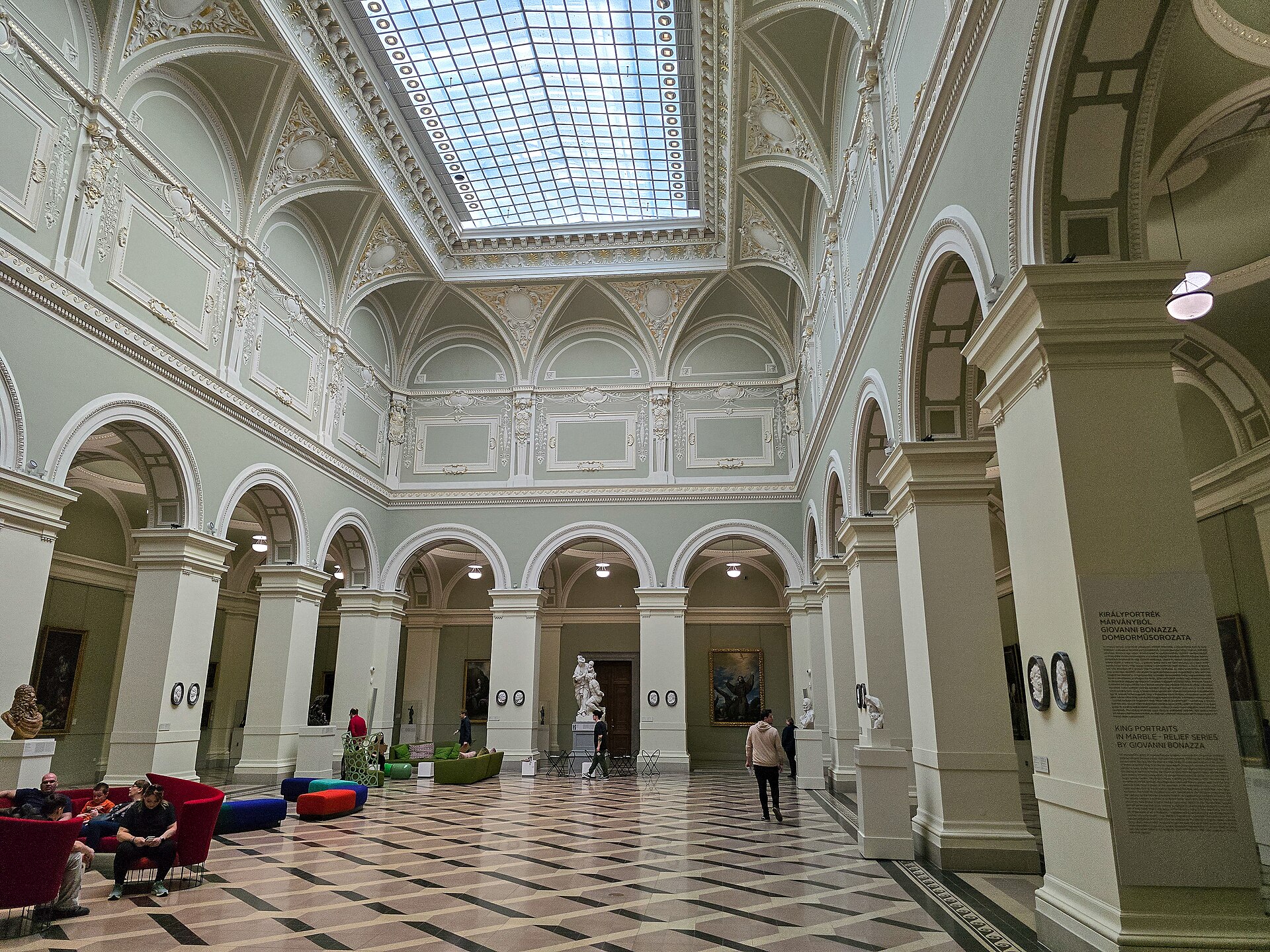 A skylit sculpture gallery with marble relief panels on the walls and statues on plinths