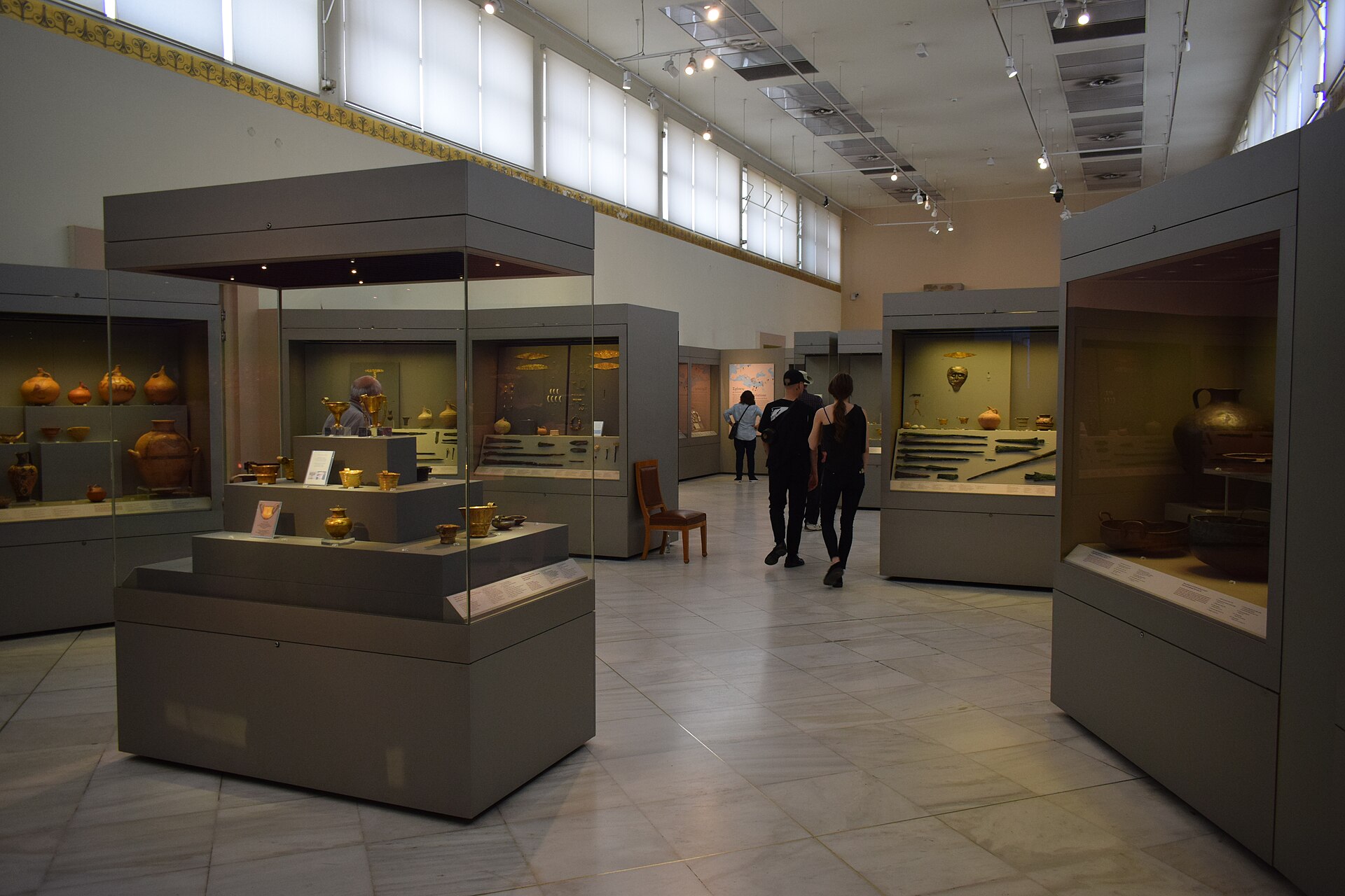 A gallery of tall display cases holding gold Mycenaean vessels and jewellery, with a pyramid-shaped central case of gold grave goods