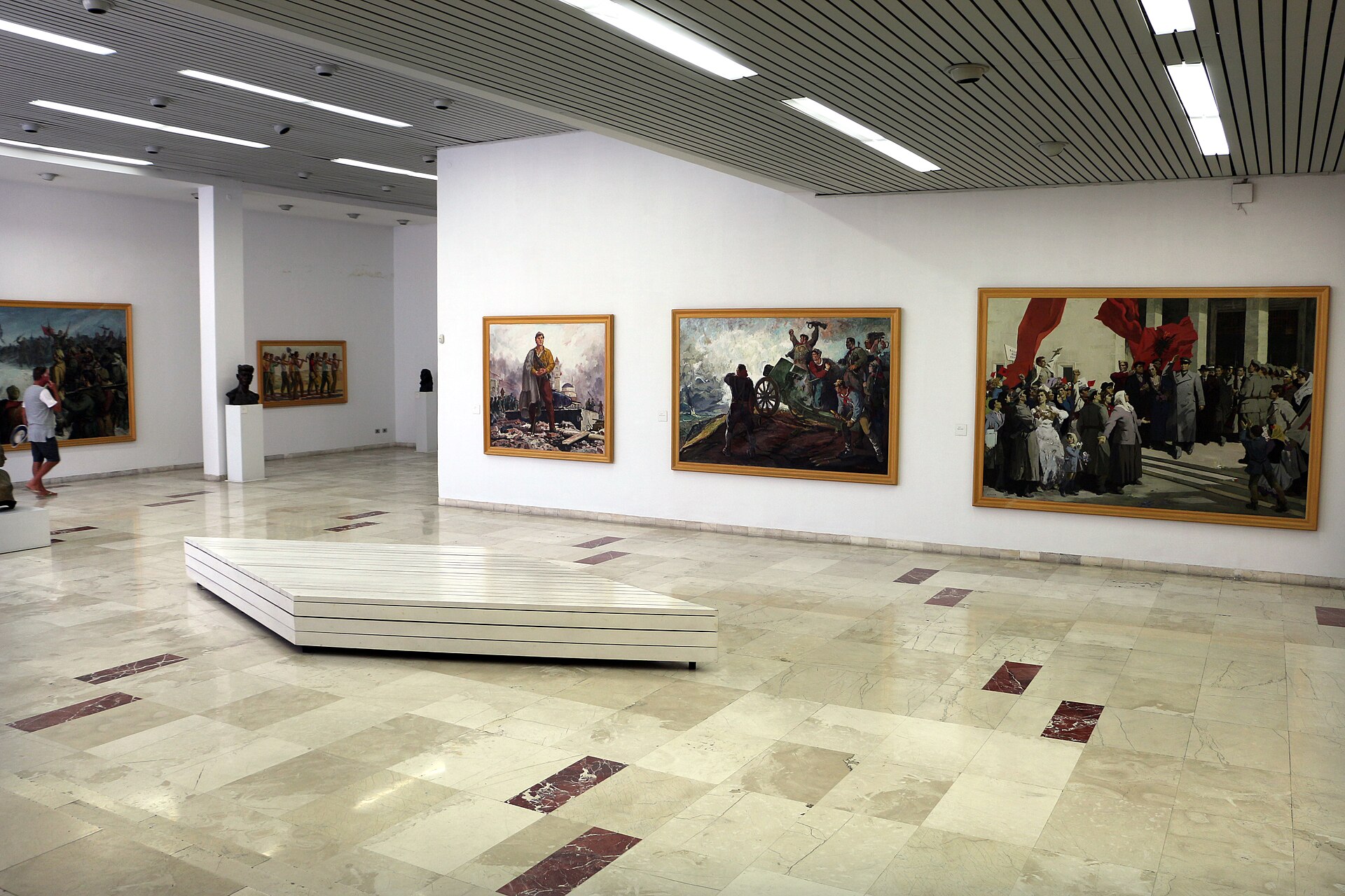A gallery room hung with large Socialist Realist paintings of partisan and civic scenes, marble floor, mid-20th-century busts on plinths