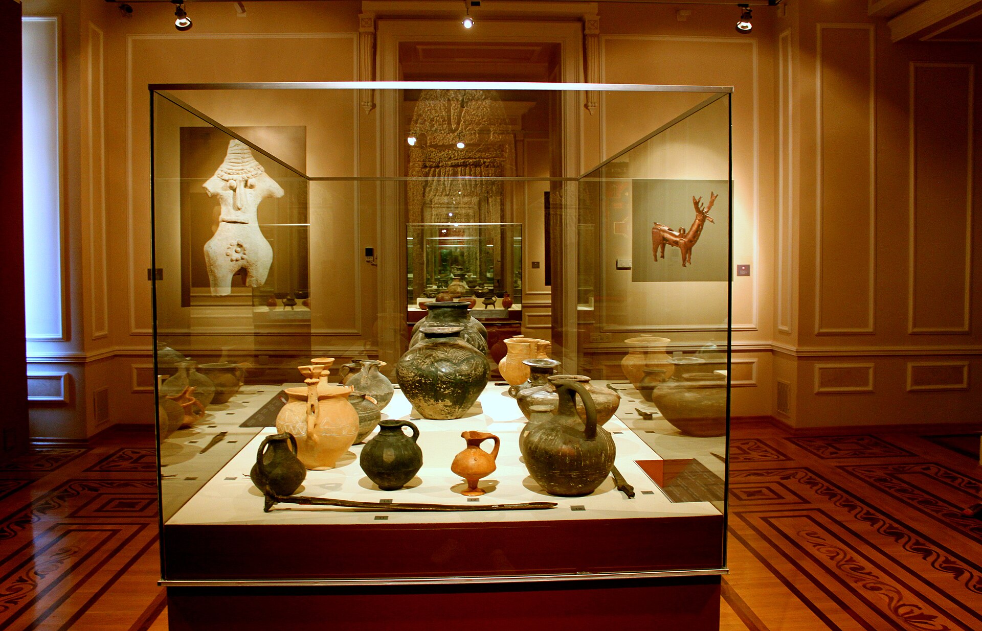A glass display case of ancient clay and bronze vessels, with a stone idol and a bronze animal figure mounted on the wall behind