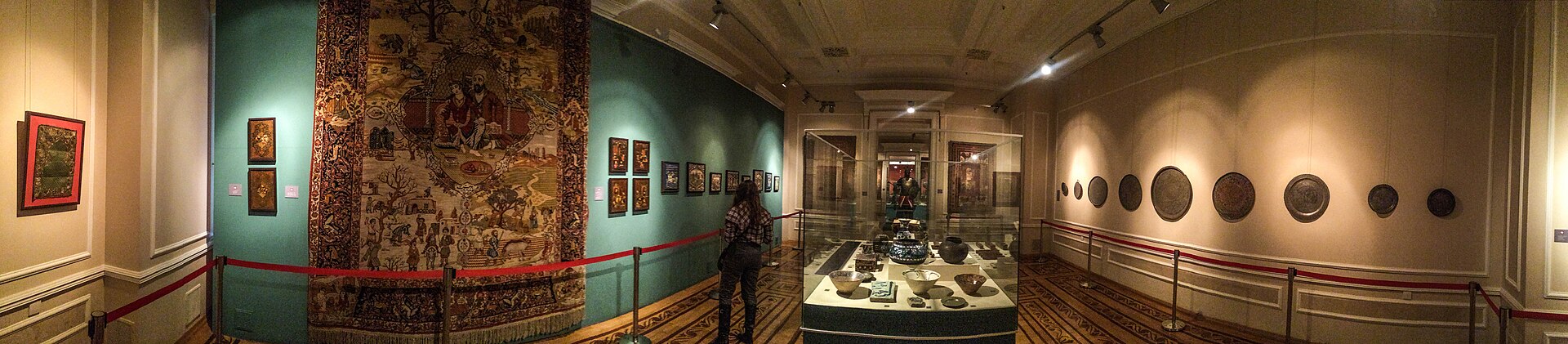 A gallery hung with a large embroidered textile and framed miniatures, with ceramics displayed in a glass case and metal plates on the wall