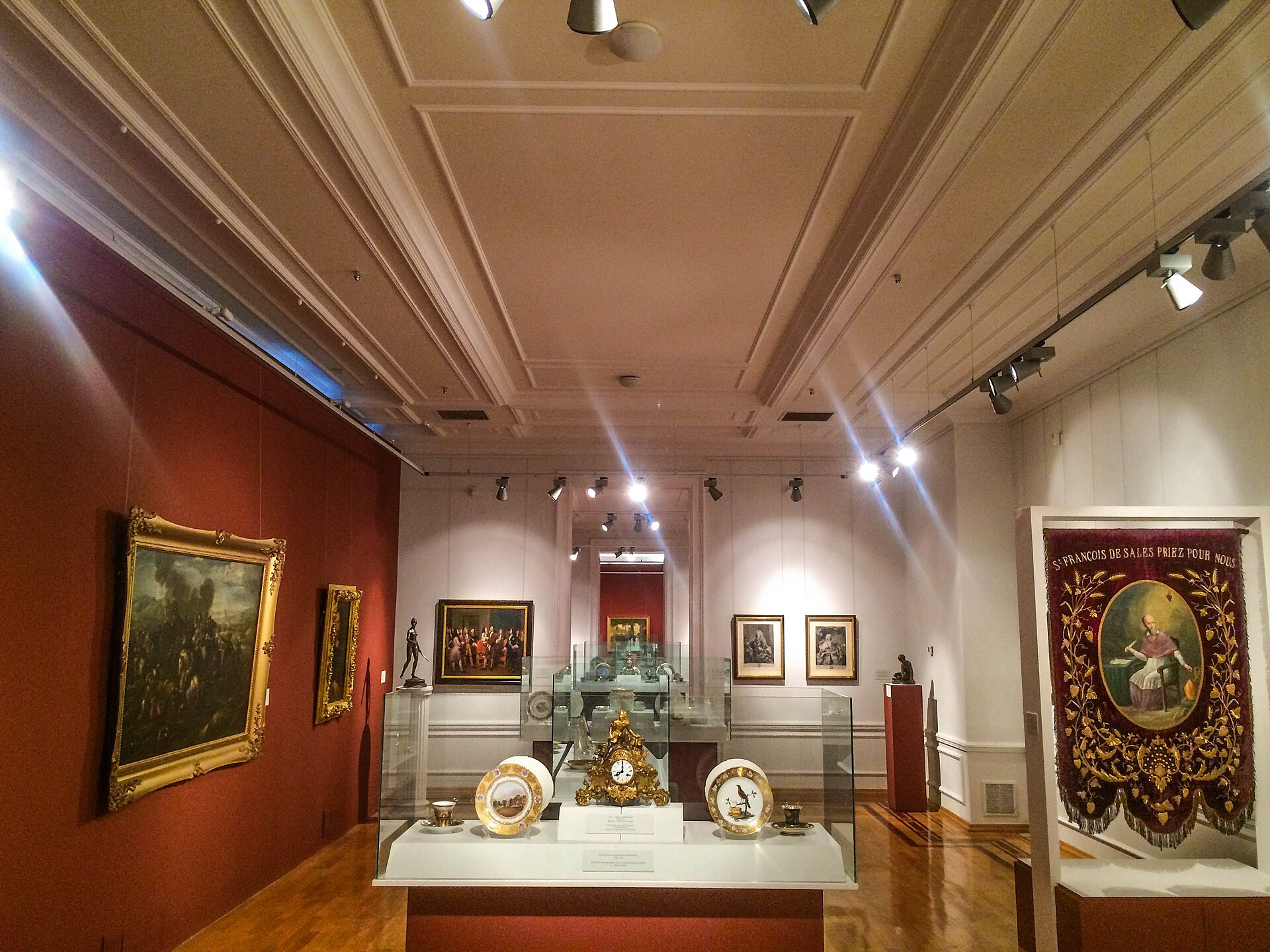 A gallery of the French art hall in the first building, red walls hung with framed paintings around glass display cases of clocks and porcelain