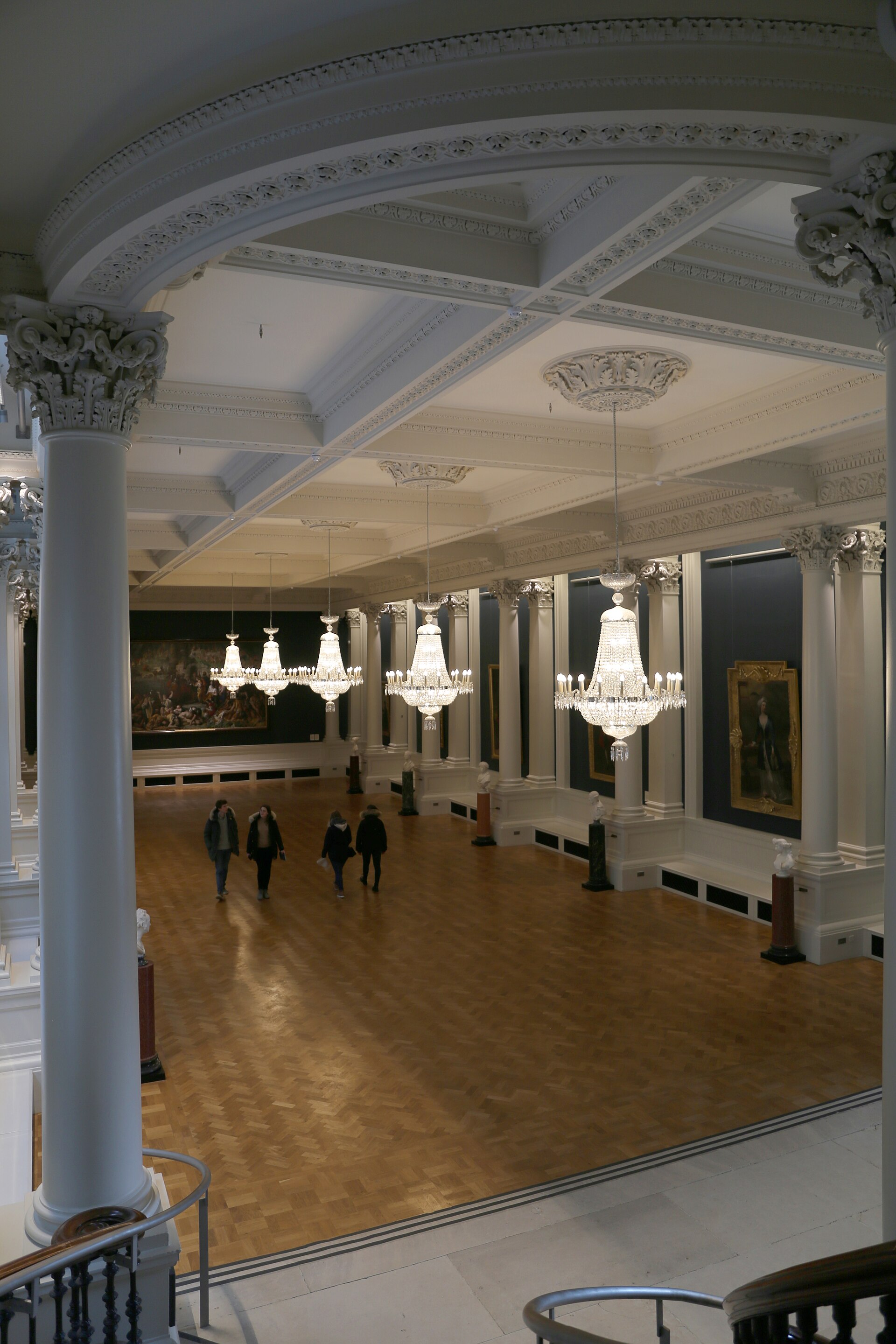 A grand colonnaded hall at entry level in the historic wing, hung with large-scale paintings beneath a gilded coffered ceiling and chandeliers