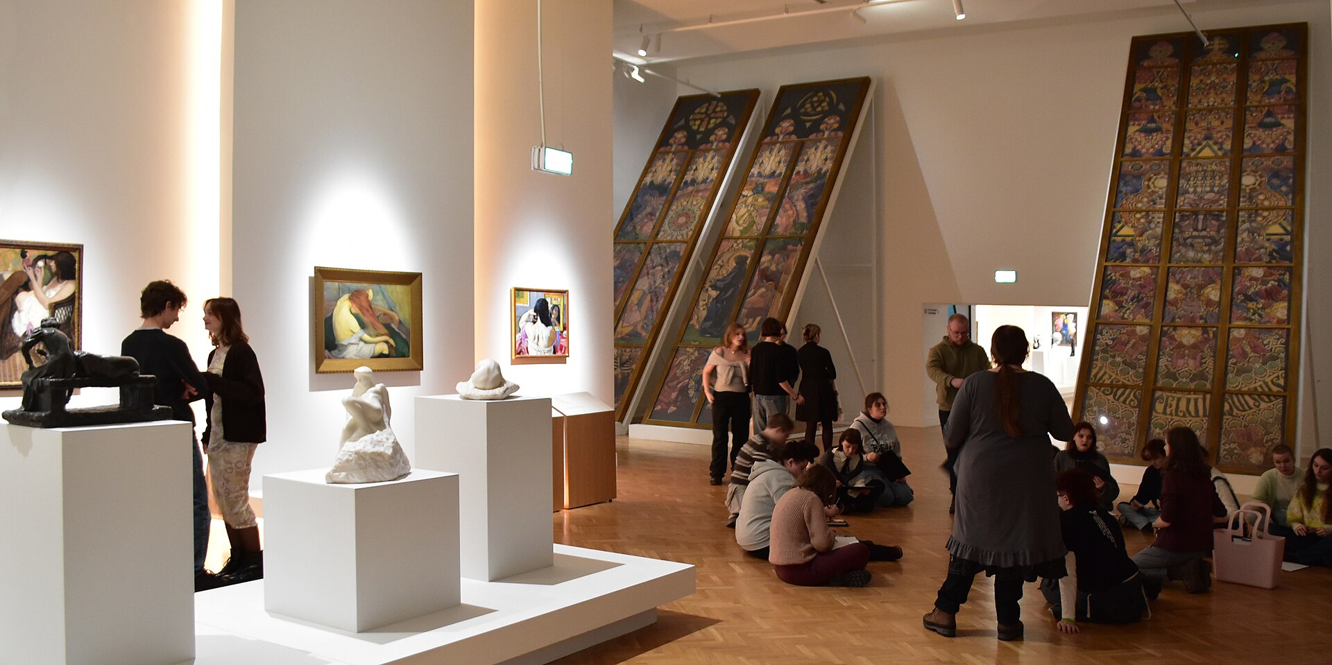 A white-walled gallery room hung with paintings, sculptures on plinths, and large textile panels, with a group of visitors seated on the parquet floor