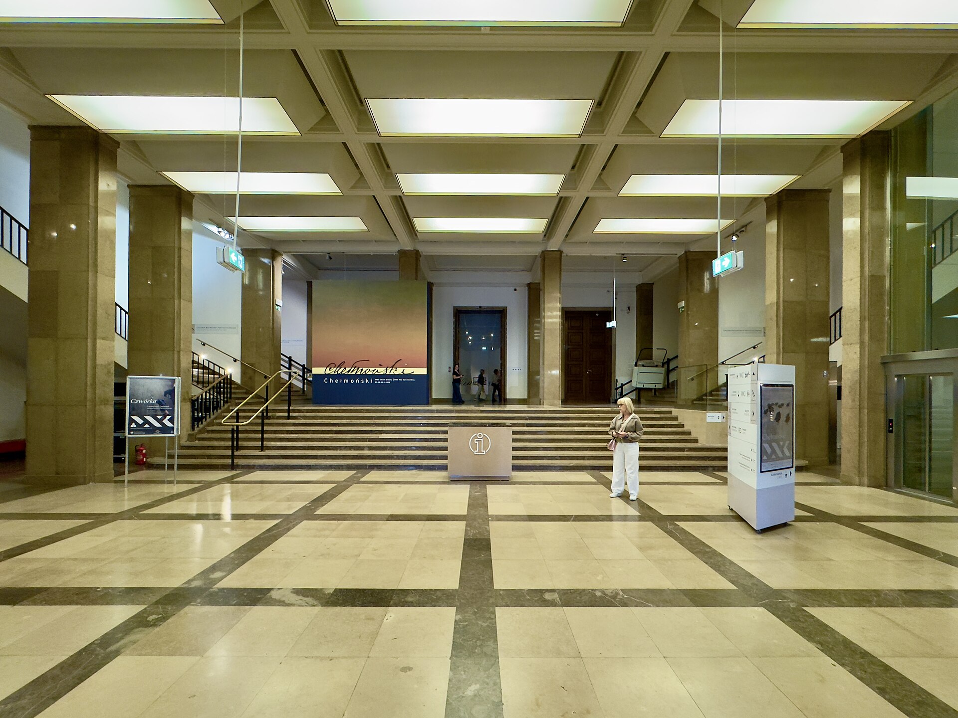 A marble-floored lobby with square stone columns, a coffered ceiling and twin staircases leading up toward an exhibition banner
