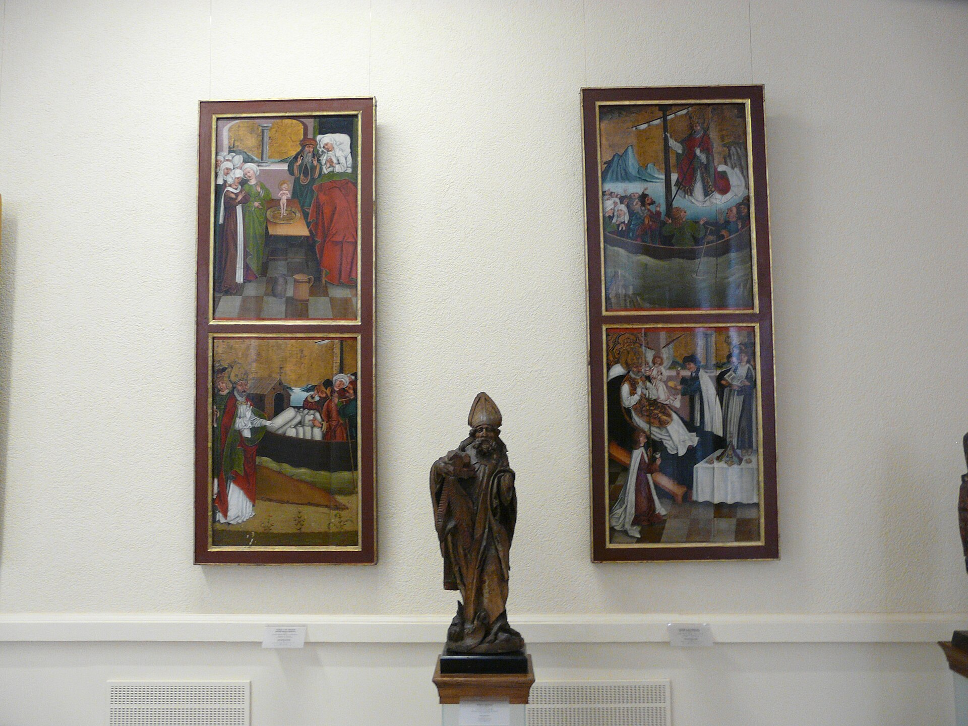 Two hinged Gothic altarpiece wings with painted narrative scenes flanking a carved wooden bishop-saint figure, in a gallery of the National Museum in Poznan