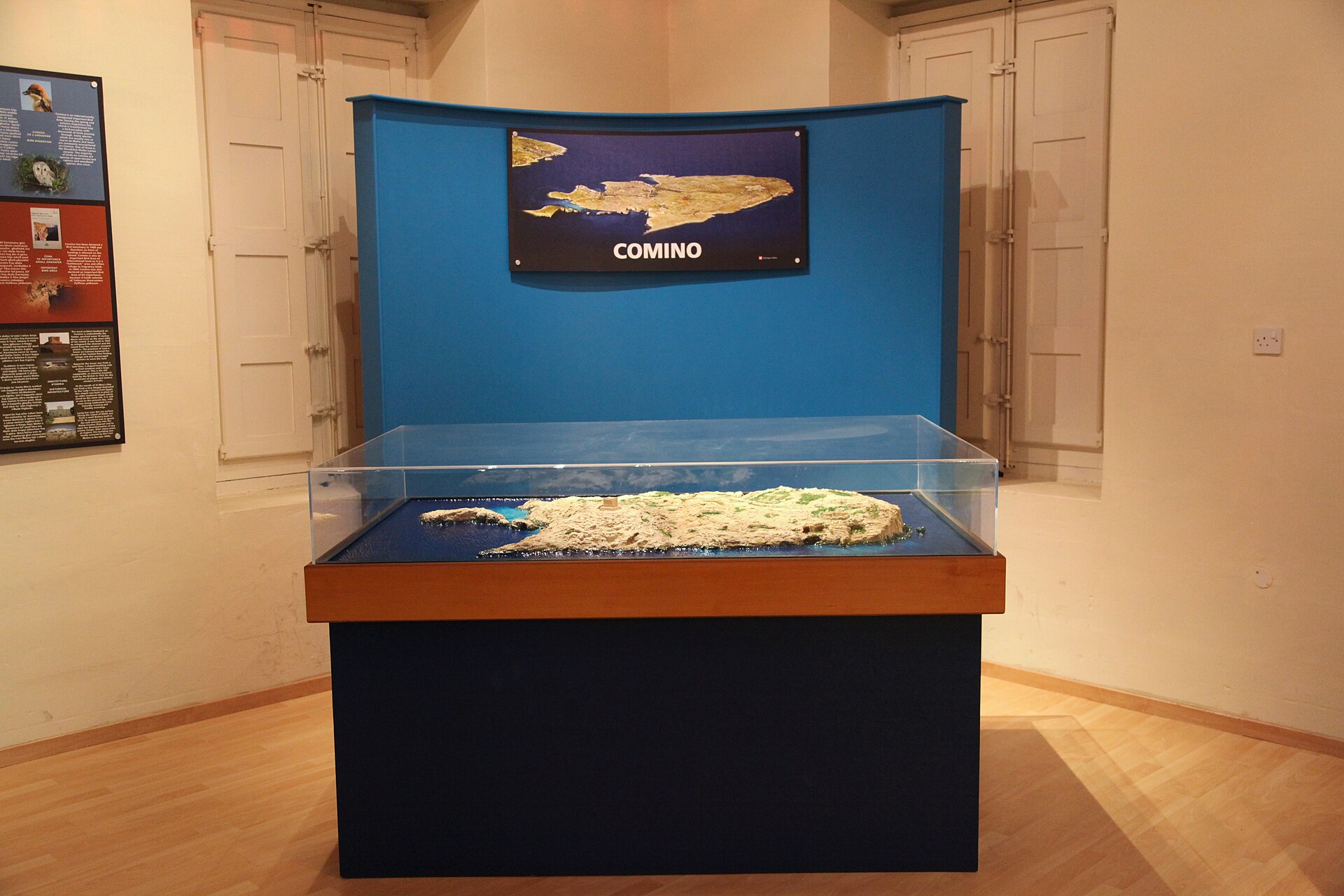 A glass display case holding a scale model of the island of Comino, with a wall panel reading COMINO