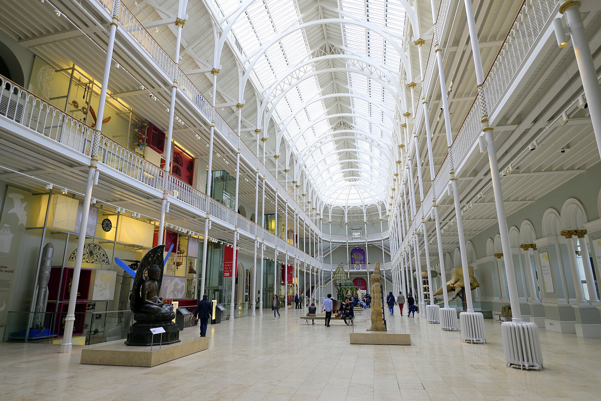 The Grand Gallery atrium, with tiers of white cast-iron columns and balustrades rising to a curved glass roof, a bronze figure and museum visitors on the tiled floor below