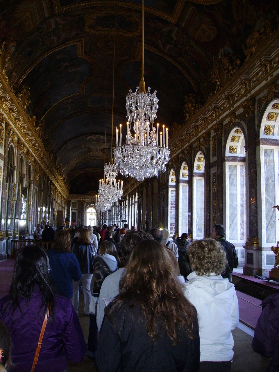The Hall of Mirrors, its arched windows and mirrored wall lit by hanging crystal chandeliers, with visitors walking its length