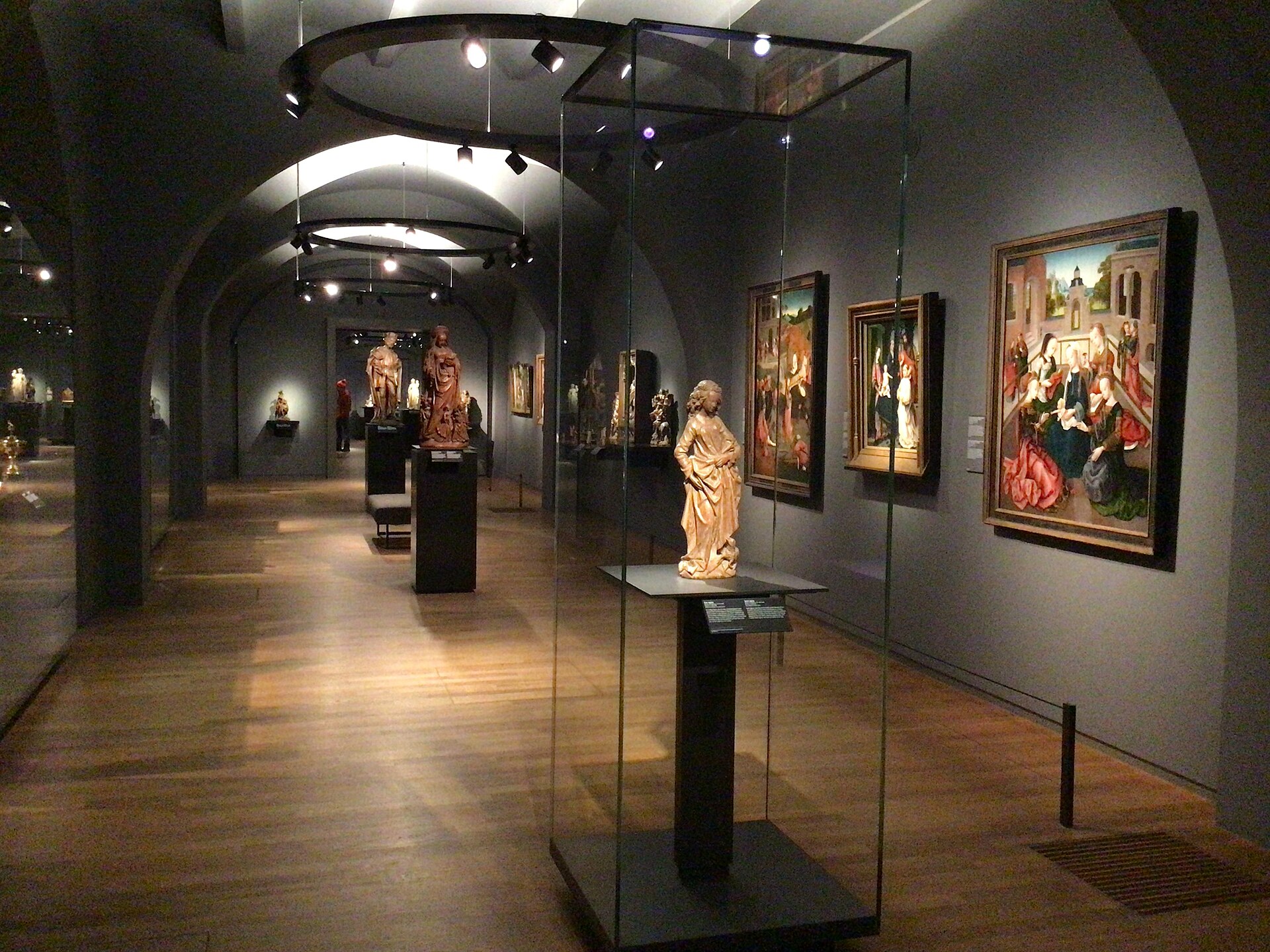 A vaulted lower-floor gallery with polychrome wood sculpture in glass cases and early Netherlandish panels on grey walls