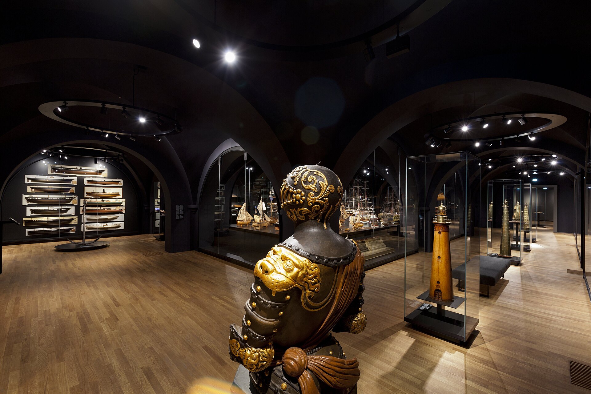 A vaulted basement room of ship models in glass cases, a gilded stern carving in the foreground