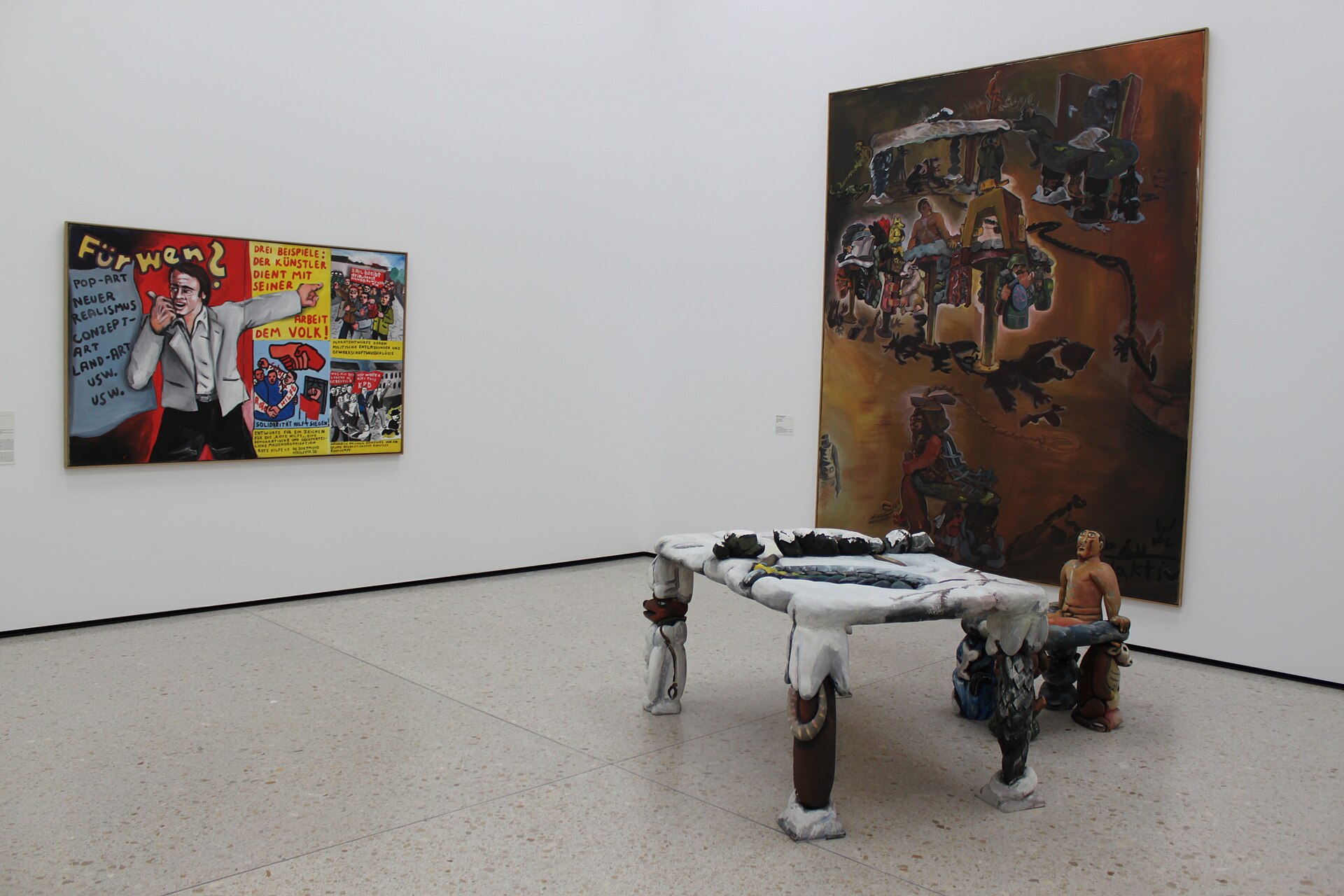 A white-walled gallery of twentieth-century paintings and a political-art collage, with a painted table sculpture in the foreground
