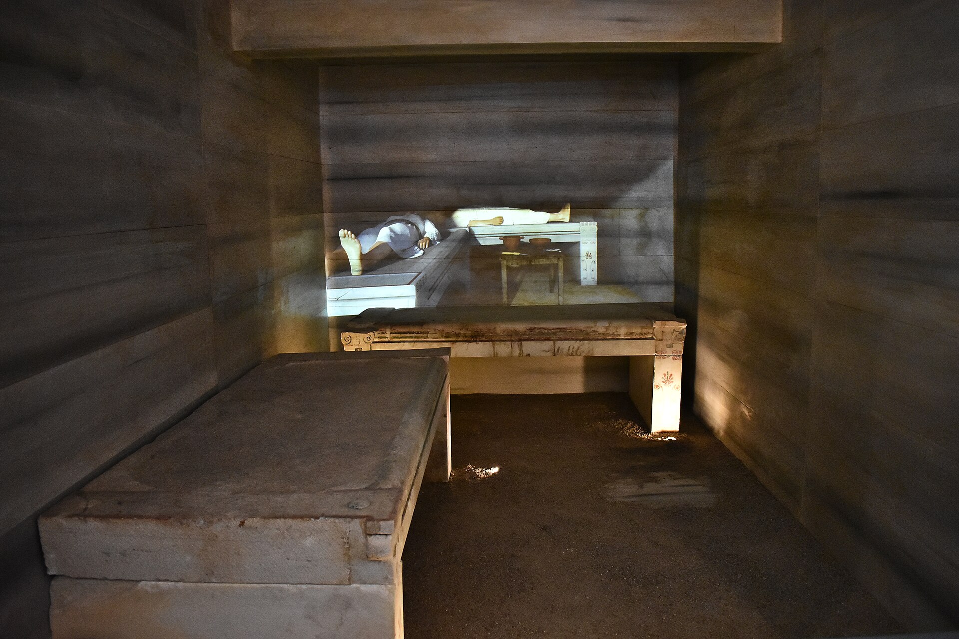 Inside the reconstructed Dedetepe tumulus burial chamber, two stone couches under a low ceiling, with a projected figure shown reclining on the far couch