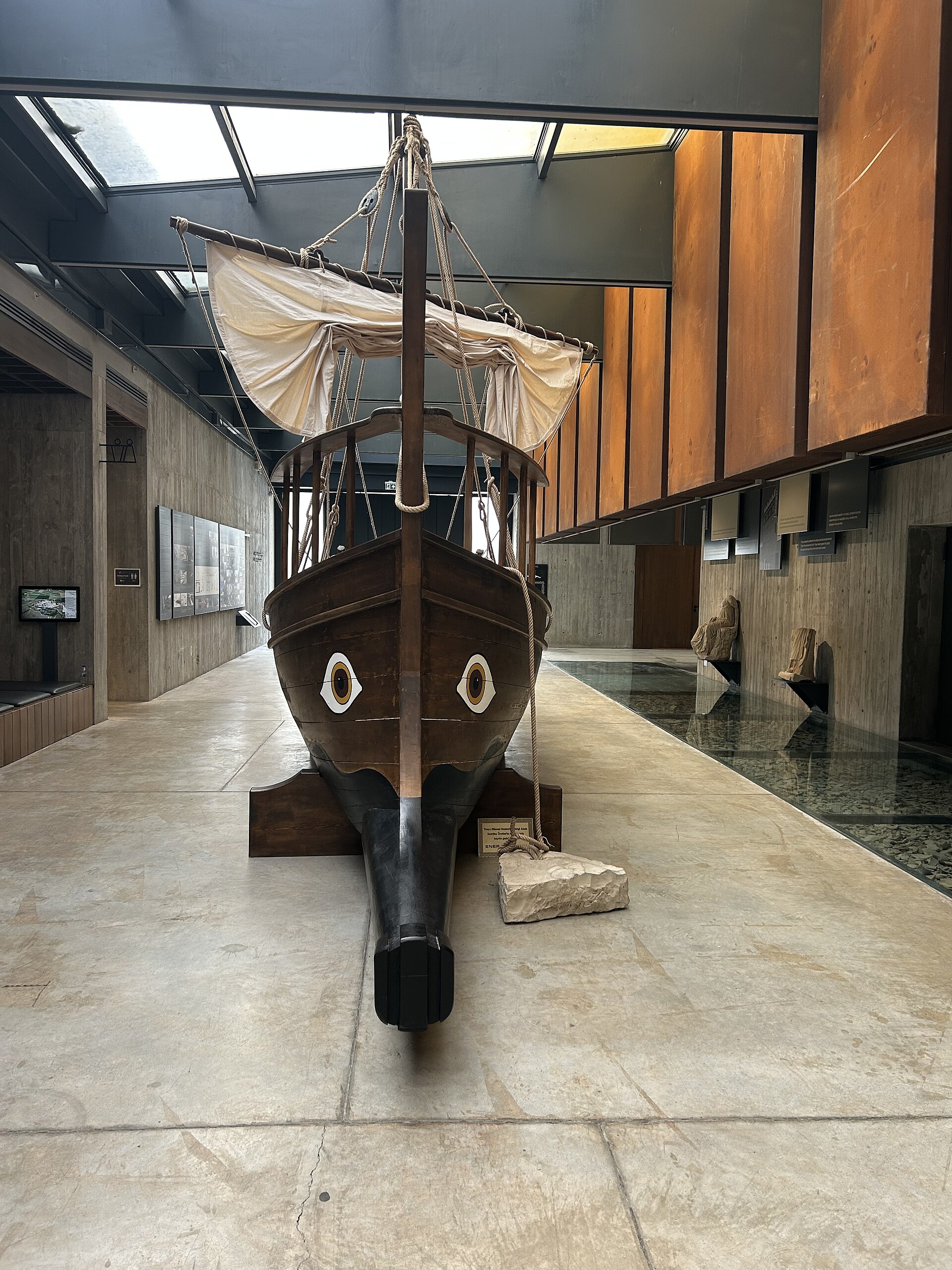 The carved wooden bow of a reconstructed sailing vessel displayed in a museum corridor, its mast and furled sail rising toward a glazed roof, with wall panels and a screen visible alongside