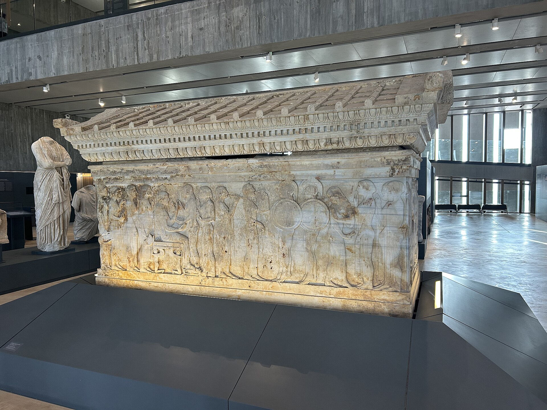 The Altikulac sarcophagus, a large carved marble coffin with a relief procession of robed figures along its long side and a pitched, dentilled lid, standing beside a draped marble statue in a gallery