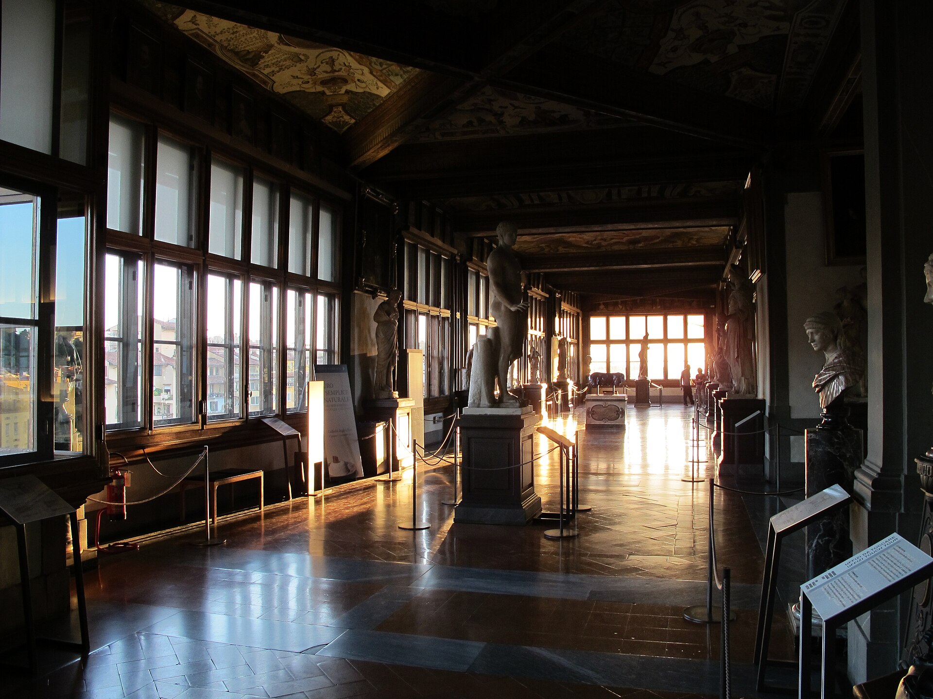 The second-floor loggia, ancient statues along a row of tall windows overlooking Florence's rooftops at sunset