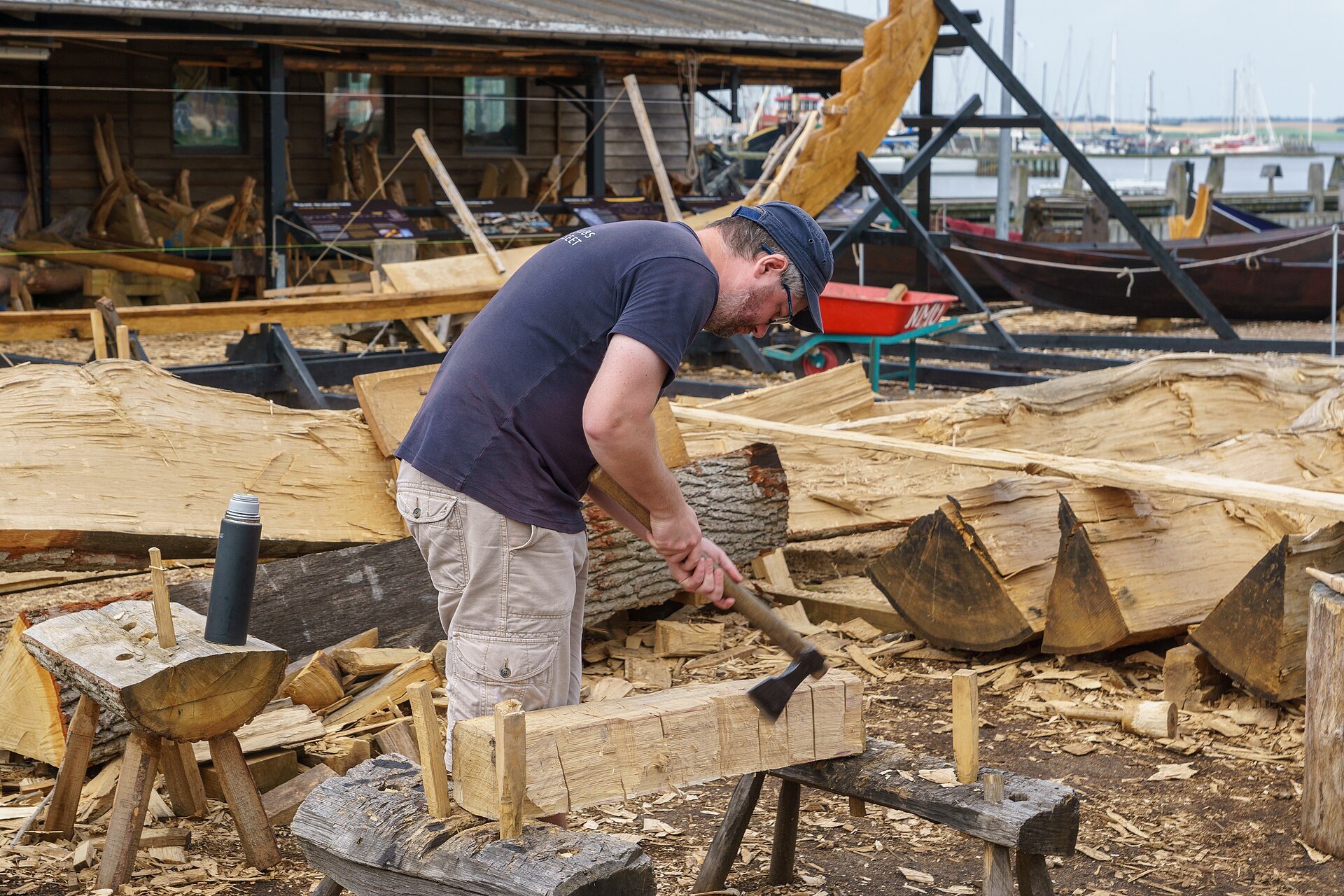 A boatbuilder splitting an oak log with an axe in the open-air boatyard, with worked timber and a half-built hull nearby